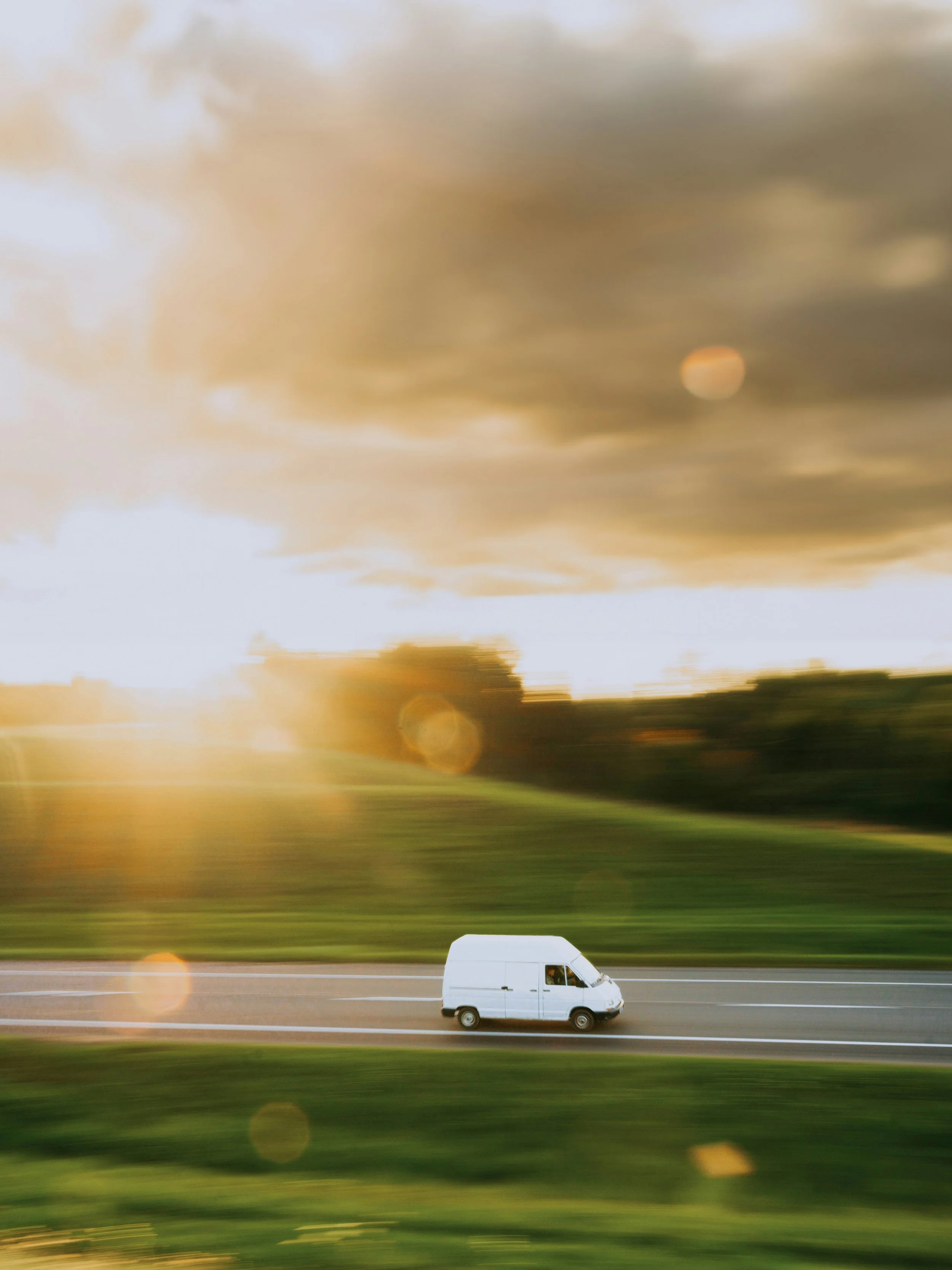 White van driving on open road during sunset with blurred background of sky and fields.