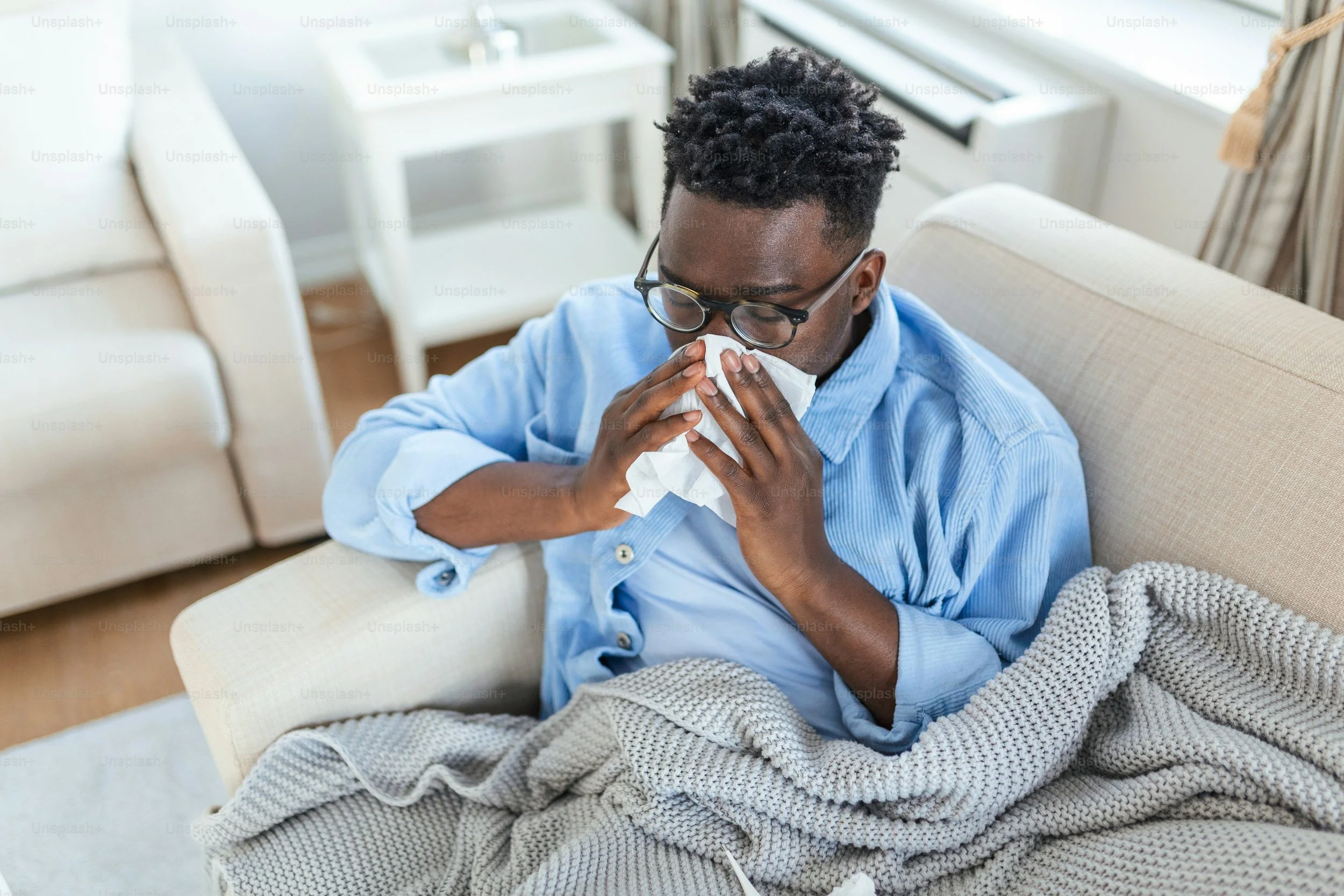 A young man with glasses and a blue shirt sitting on a beige sofa, using a tissue to blow his nose, covered with a grey knitted blanket in a bright living room.